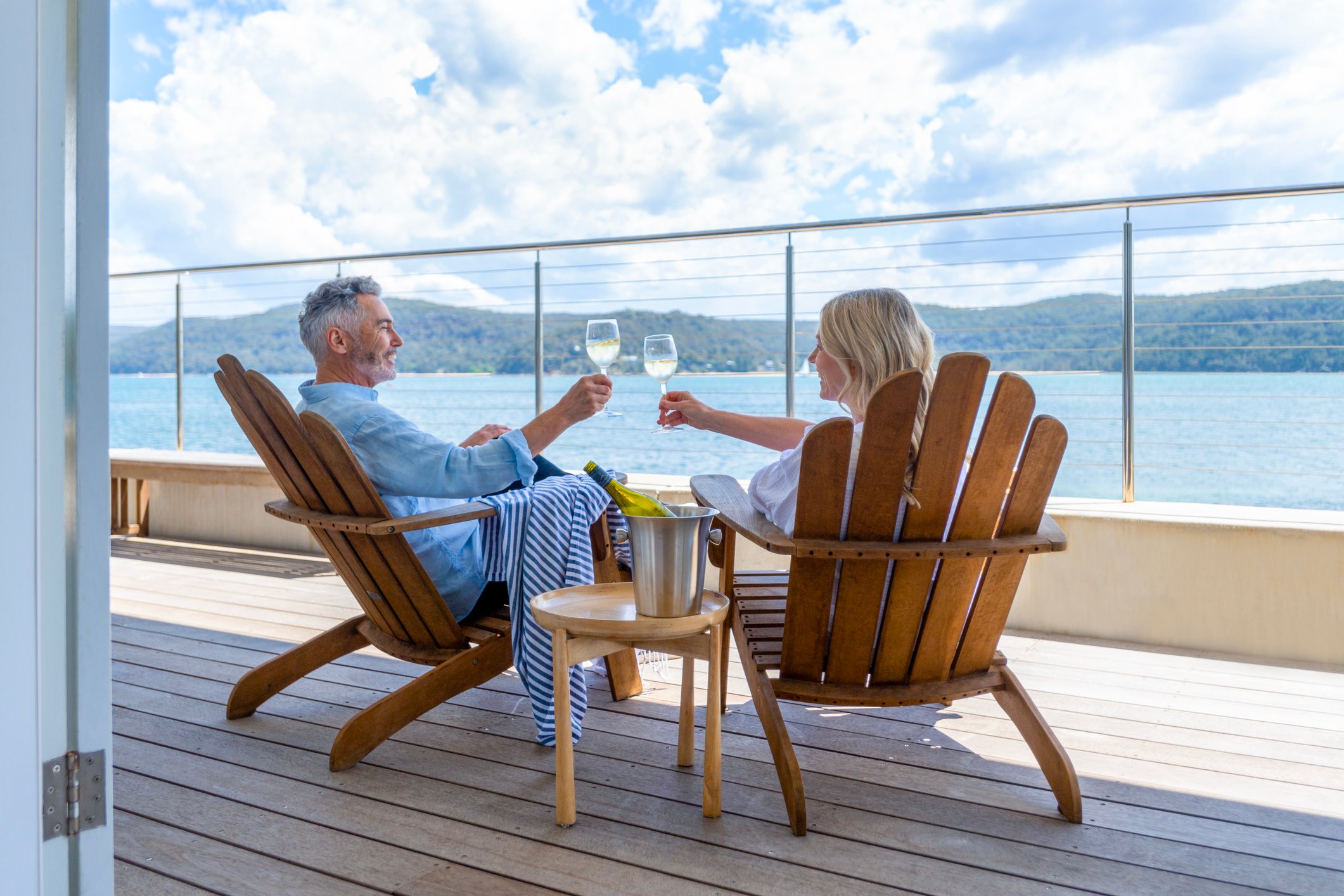 Two people toasting with glasses of wine in large wooden chairs overlooking a lake.