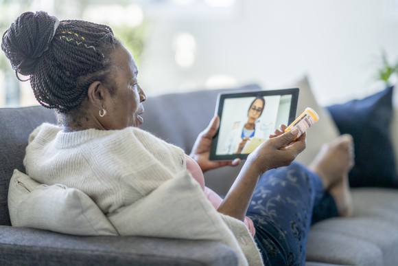 A person sits on a couch and holds a bottle of pills while talking to a healthcare professional on a tablet.