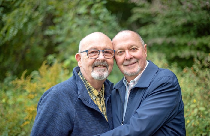 A couple is standing outside, against some trees, smiling. 