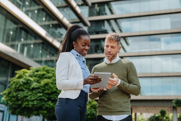 Two professionally dressed people have a discussion outside an office while looking at a tablet.