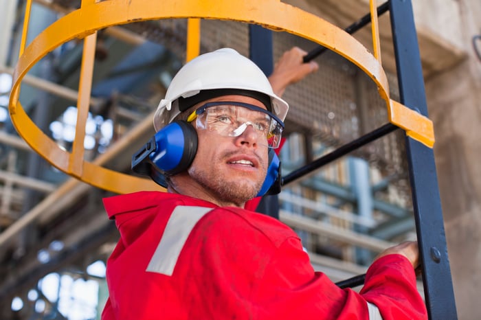 A worker wearing personal protective equipment climbs a ladder at an oil refinery.