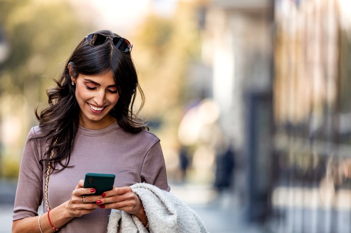 A smiling person walking and looking at a smartphone.