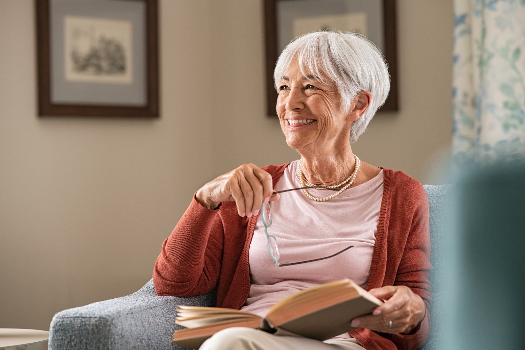 A smiling person in a chair holding eyeglasses and a book.