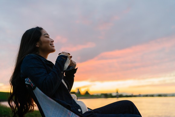 A person sips a hot beverage by a lake at sunset.