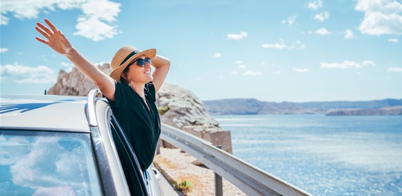 A person wearing sunglasses smiles while leaning out of a car window by the ocean.