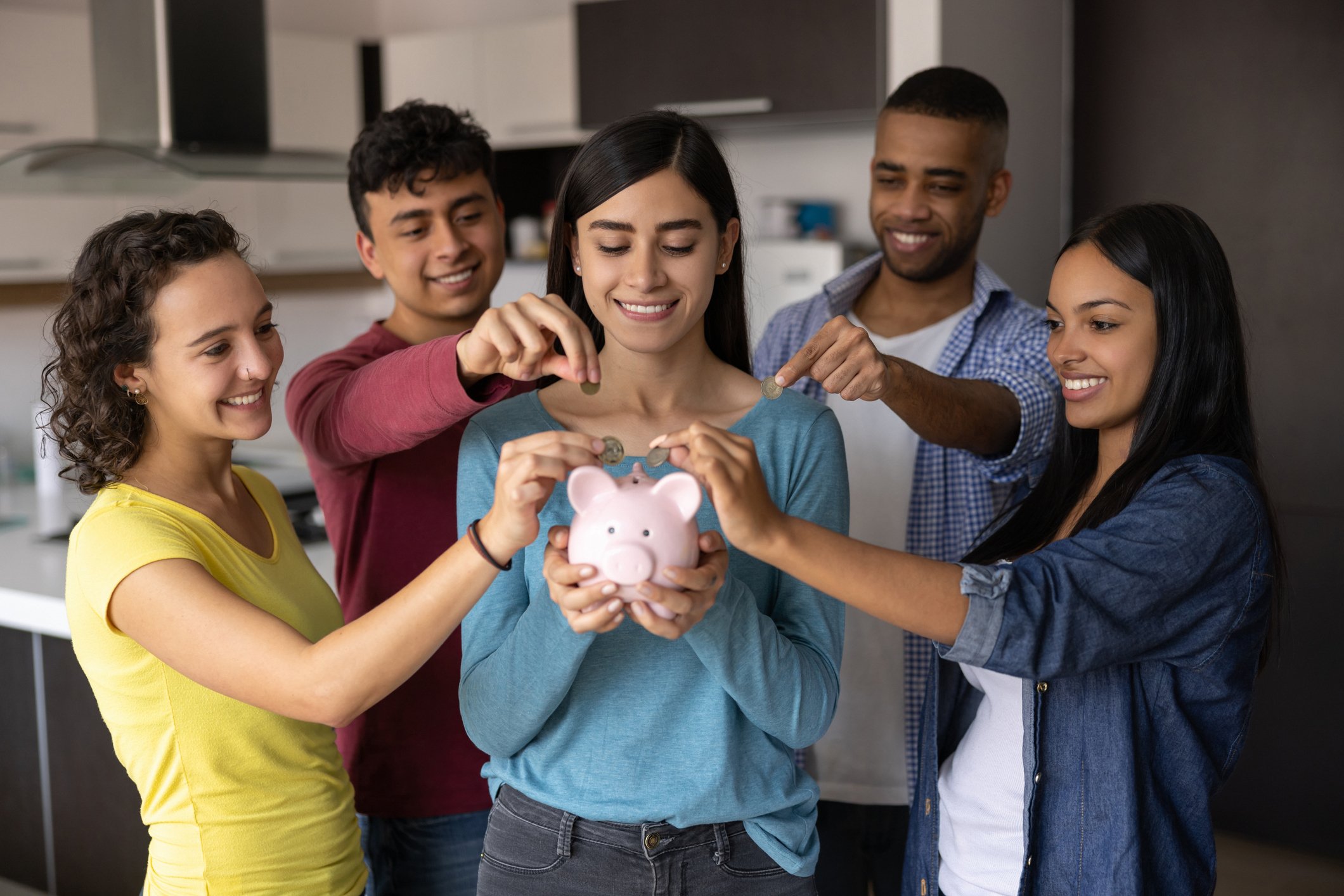 Four people surrounding a fifth and putting money in a piggy bank.