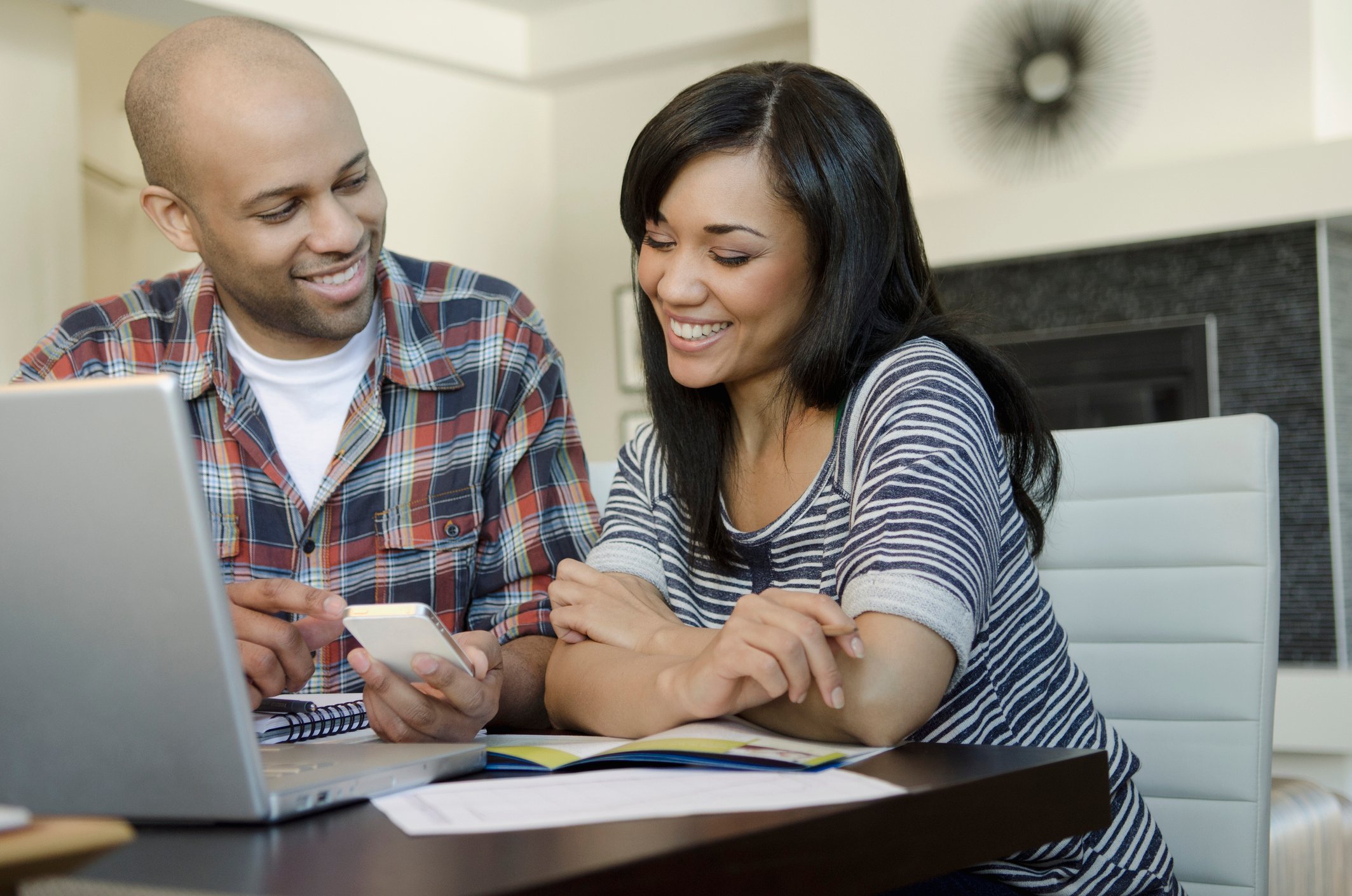 Two people smile while sitting at a table with a laptop and looking at documents. 