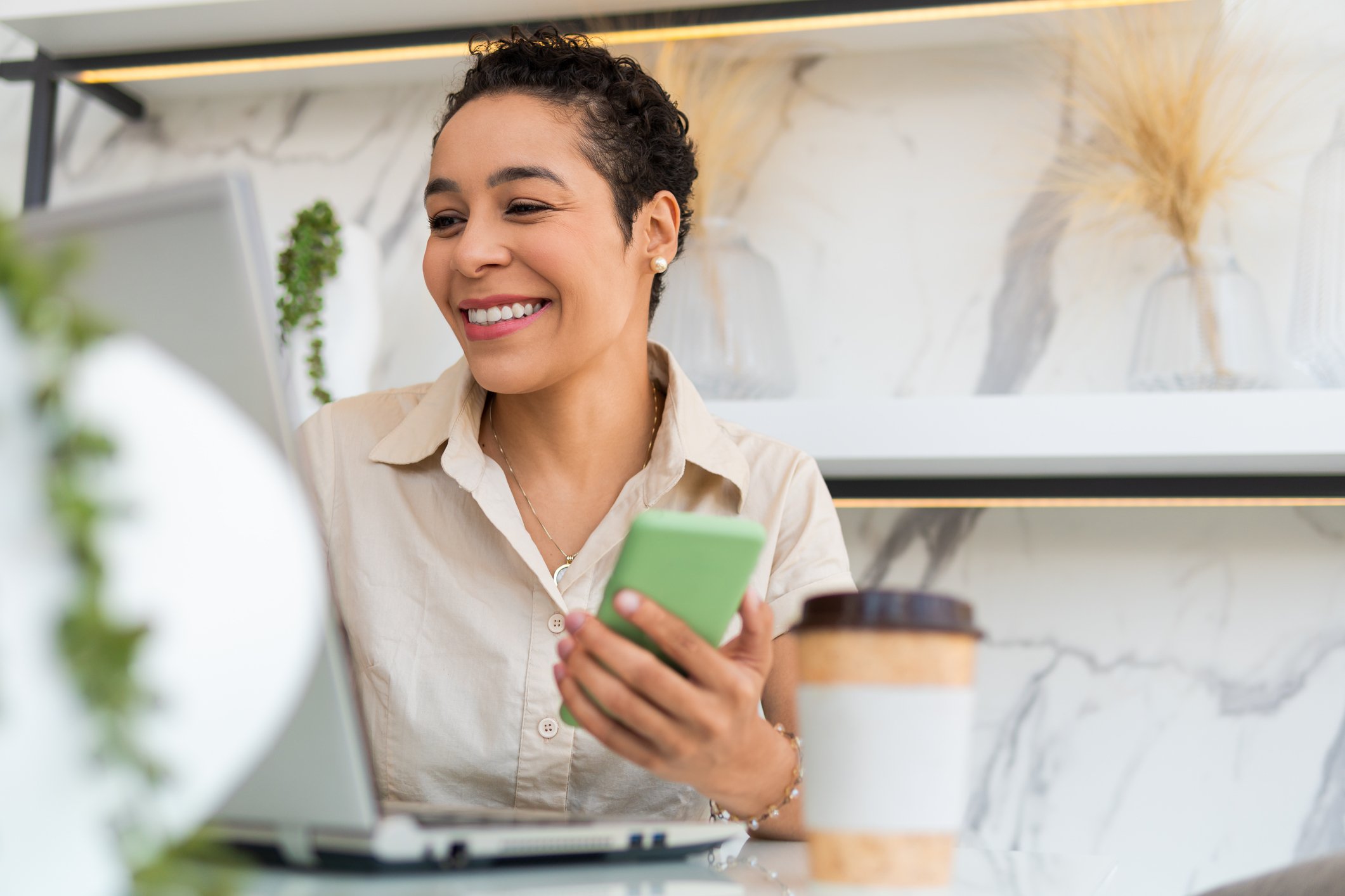 A smiling person working on a laptop and looking at a smartphone, with a coffee cup in the foreground.