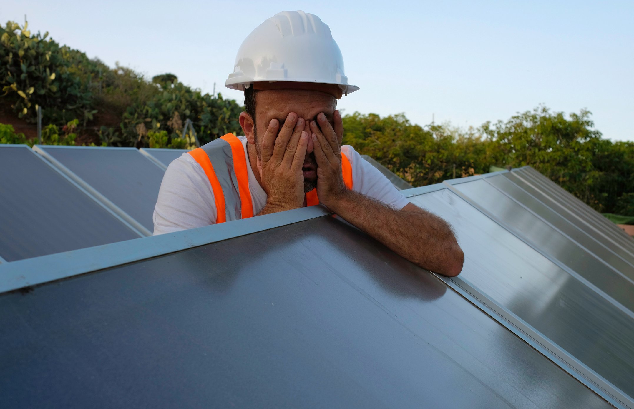 A person wearing personal protective equipment covers their face while leaning on a solar panel. 