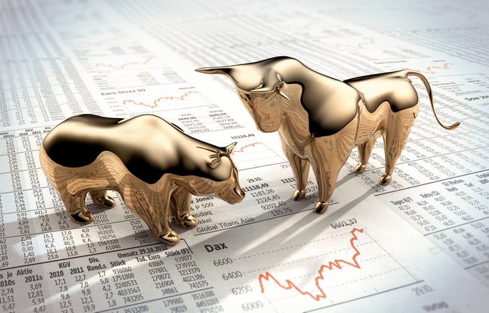 A bull figurine looks down at a bear, both standing on financial pages.