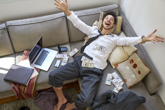 A man cheers as he lies back on his couch near piles of money.