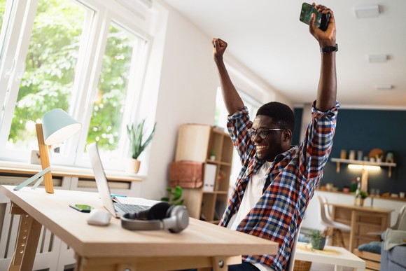 A person raises their hands in the air in celebration while sitting at a desk in front of a computer. 