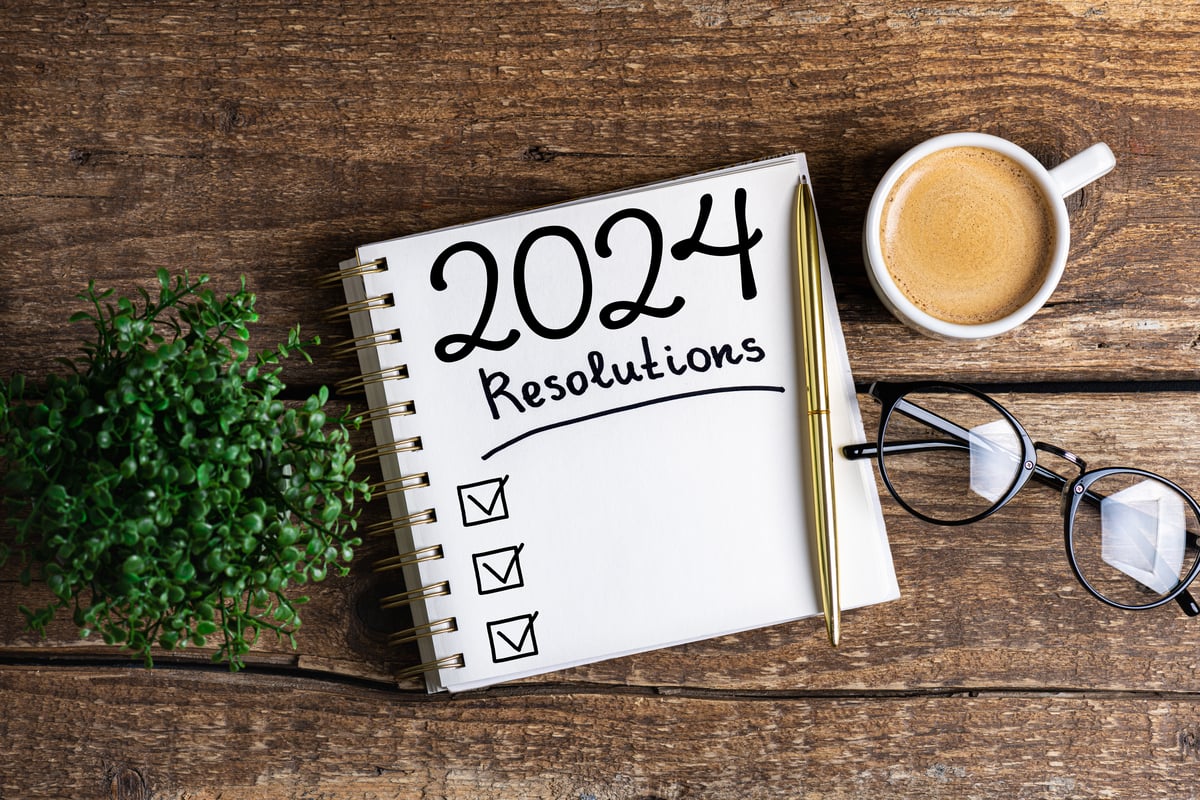 A notepad with "2024 resolutions" written on it is positioned on a table next to a cup of coffee and a pair of glasses.