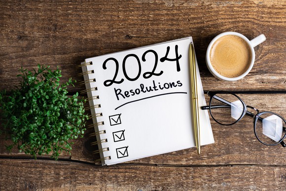 A notepad with "2024 resolutions" written on it is positioned on a table next to a cup of coffee and a pair of glasses.