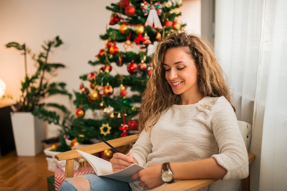 An investor sitting in front of a Christmas tree takes notes in a notebook.