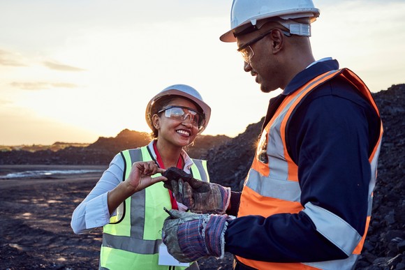 Two people wearing personal protective equipment smile at each other at a work site. 