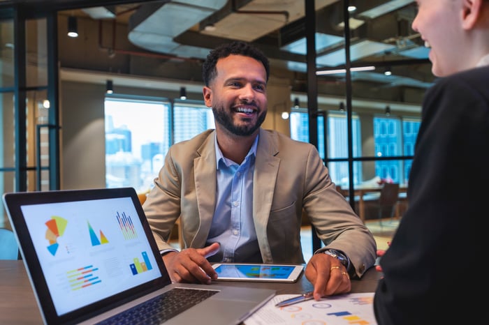 Two smiling people at a table with a laptop and tablet.