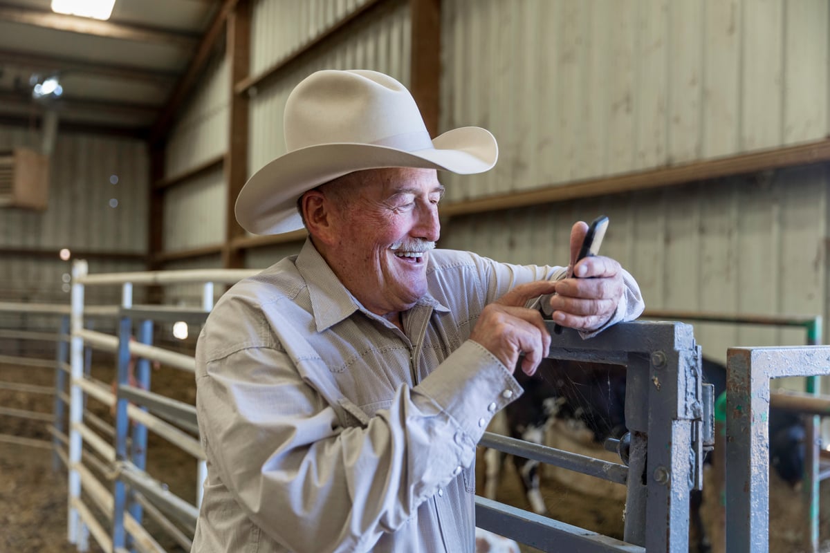 A person wearing a cowboy hat leans on a stable railing while smiling and looking at their phone. 