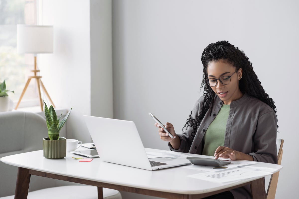 Person at desk with laptop, looking at calculator and phone. 