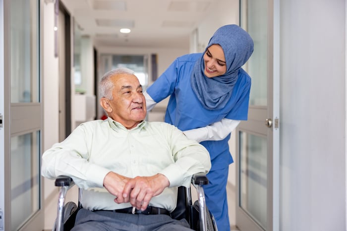 A person in scrubs pushing a person in a wheelchair.