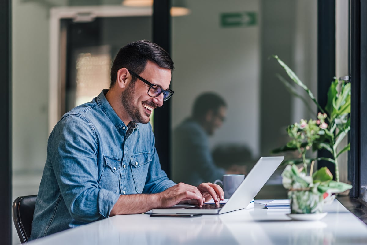 A person smiling while sitting at a desk and typing on a laptop computer.