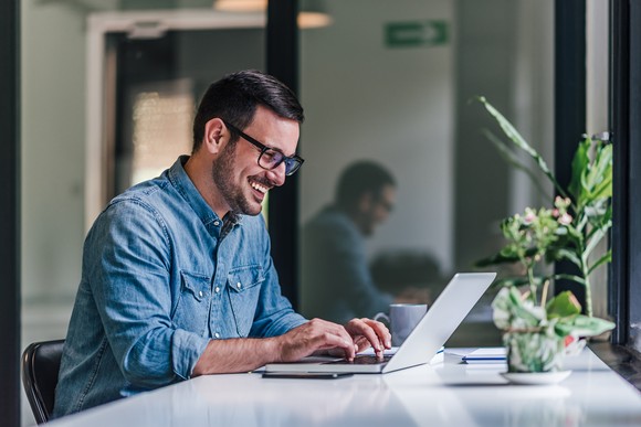 A person smiling while sitting at a desk and typing on a laptop computer.