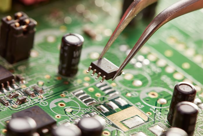 A pair of tweezers holding a computer chip over a circuit board.