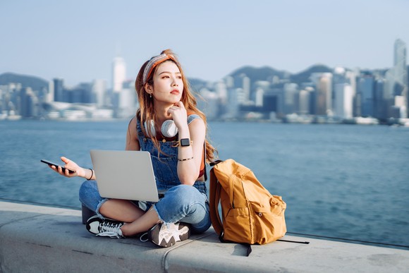 A person sitting on a wall with a laptop in front of them and Hong Kong in the background.