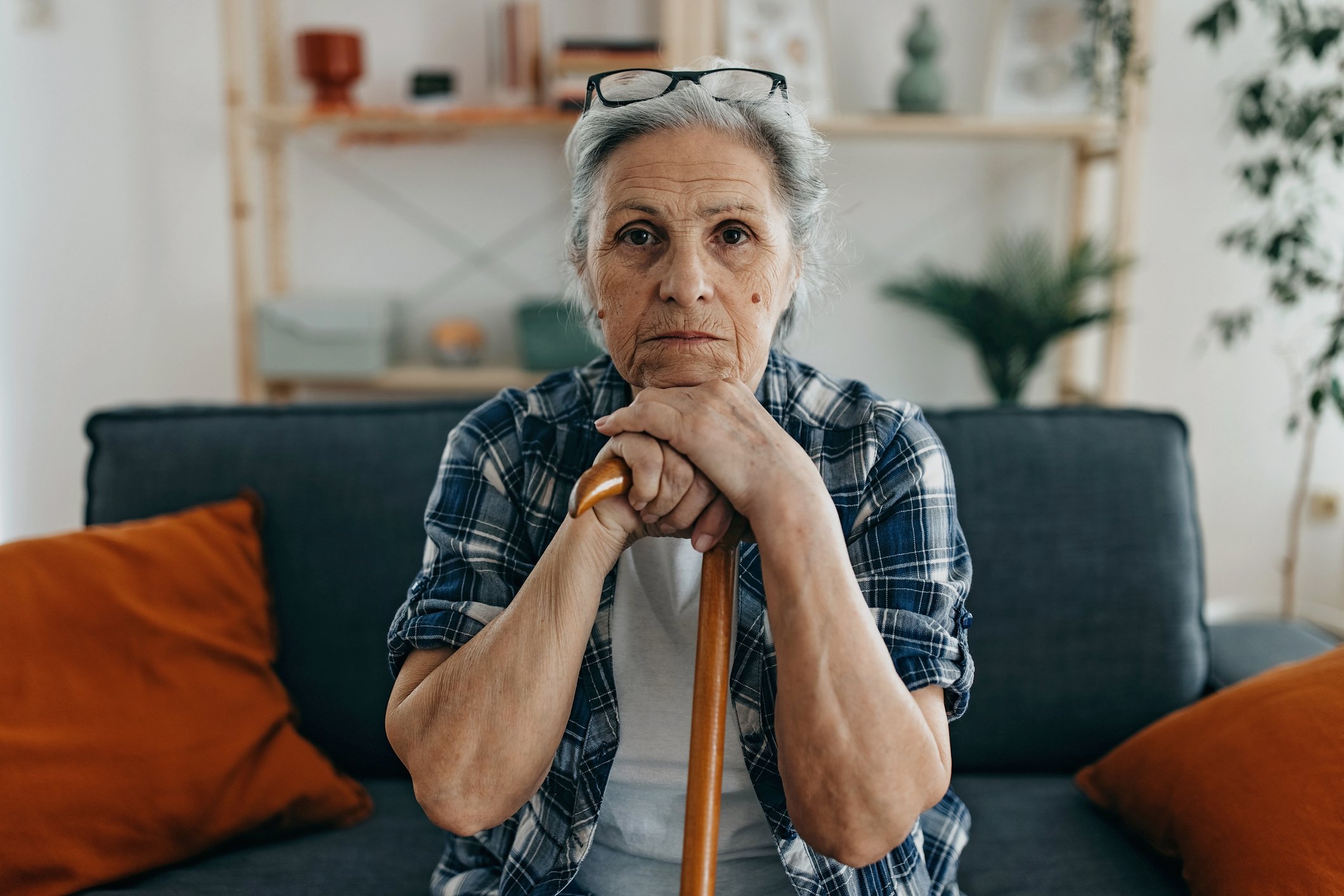 An older person sitting at home is leaning on a cane and looking very serious. 