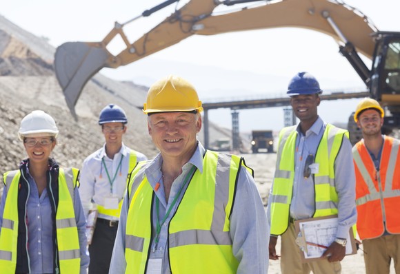 Construction workers standing in front of a backhoe. 