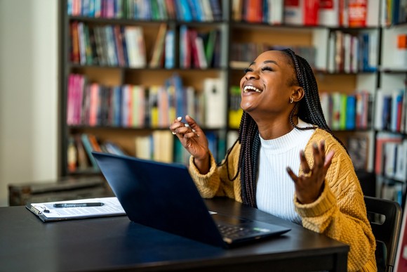 A person smiles while looking up and sitting at a desk in a library in front of a laptop computer.