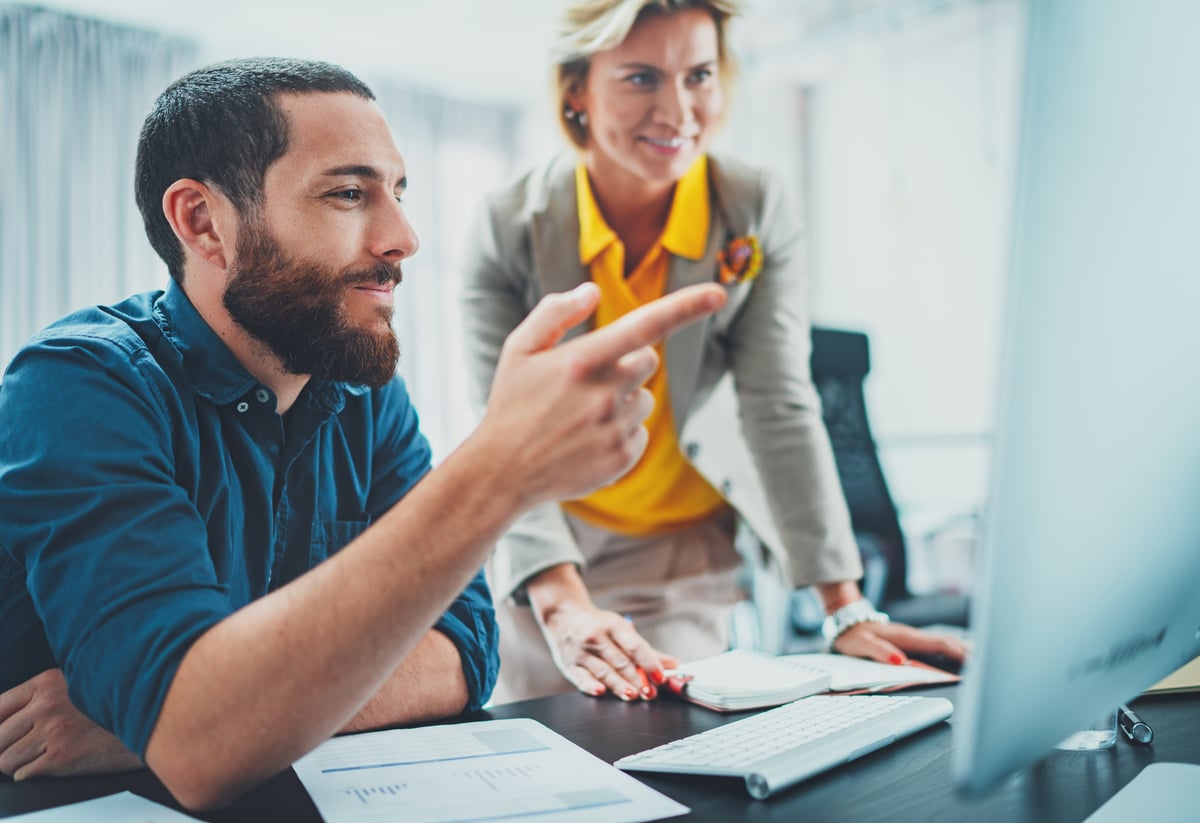 A seated, smiling person points out something on a computer screen to a standing, smiling person.
