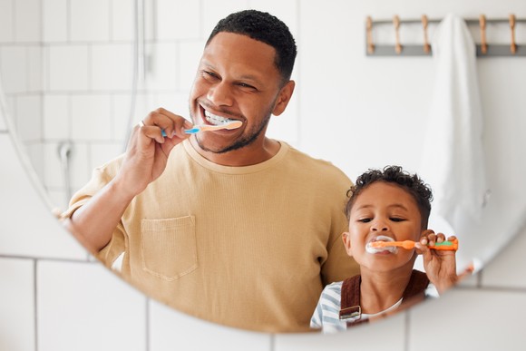 An adult and child brush their teeth while looking in a mirror in a bathroom.