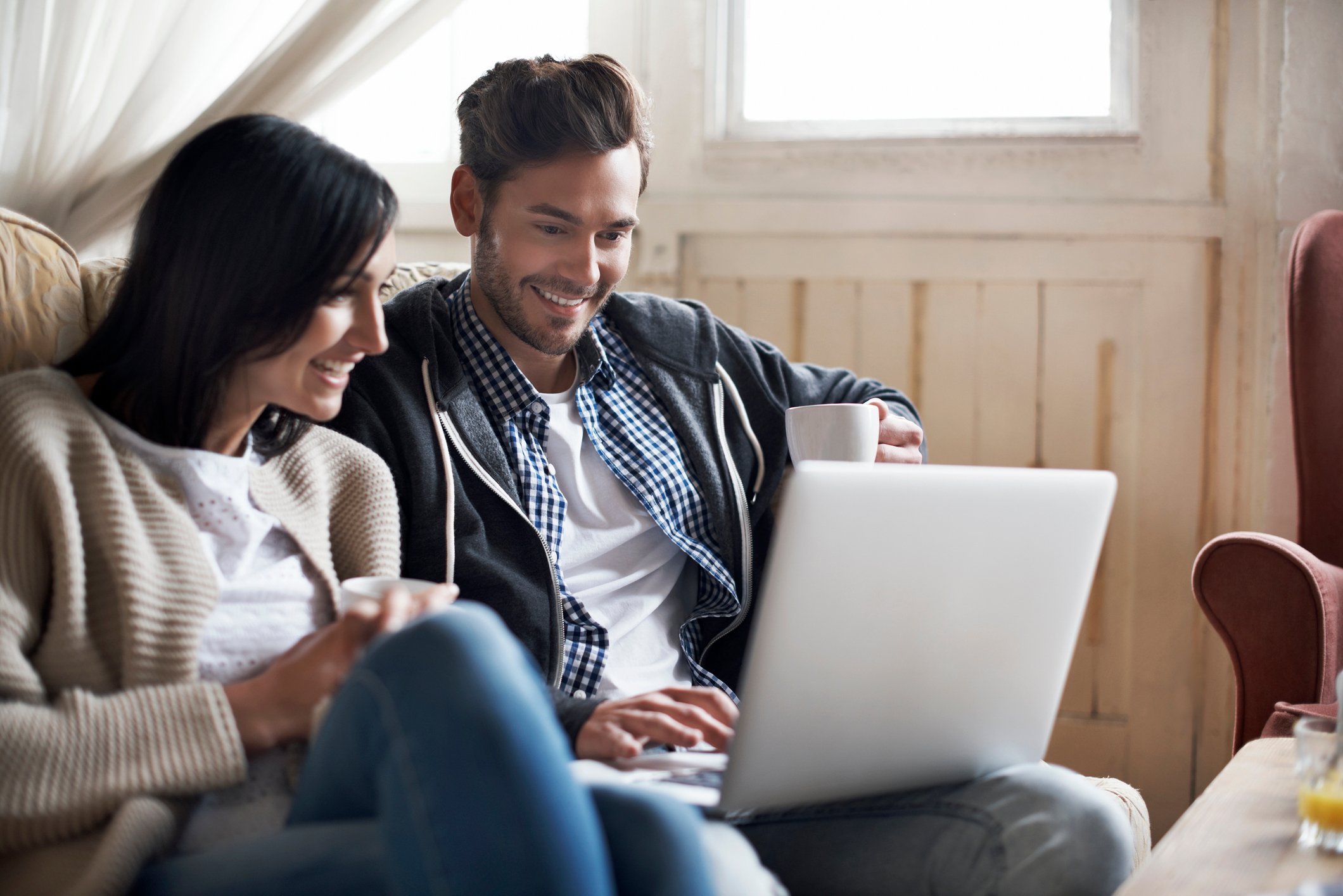 Two people sitting on a couch smile while looking at something on a laptop.