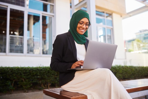 A person sitting on a bench smiles while interacting with a laptop computer. 