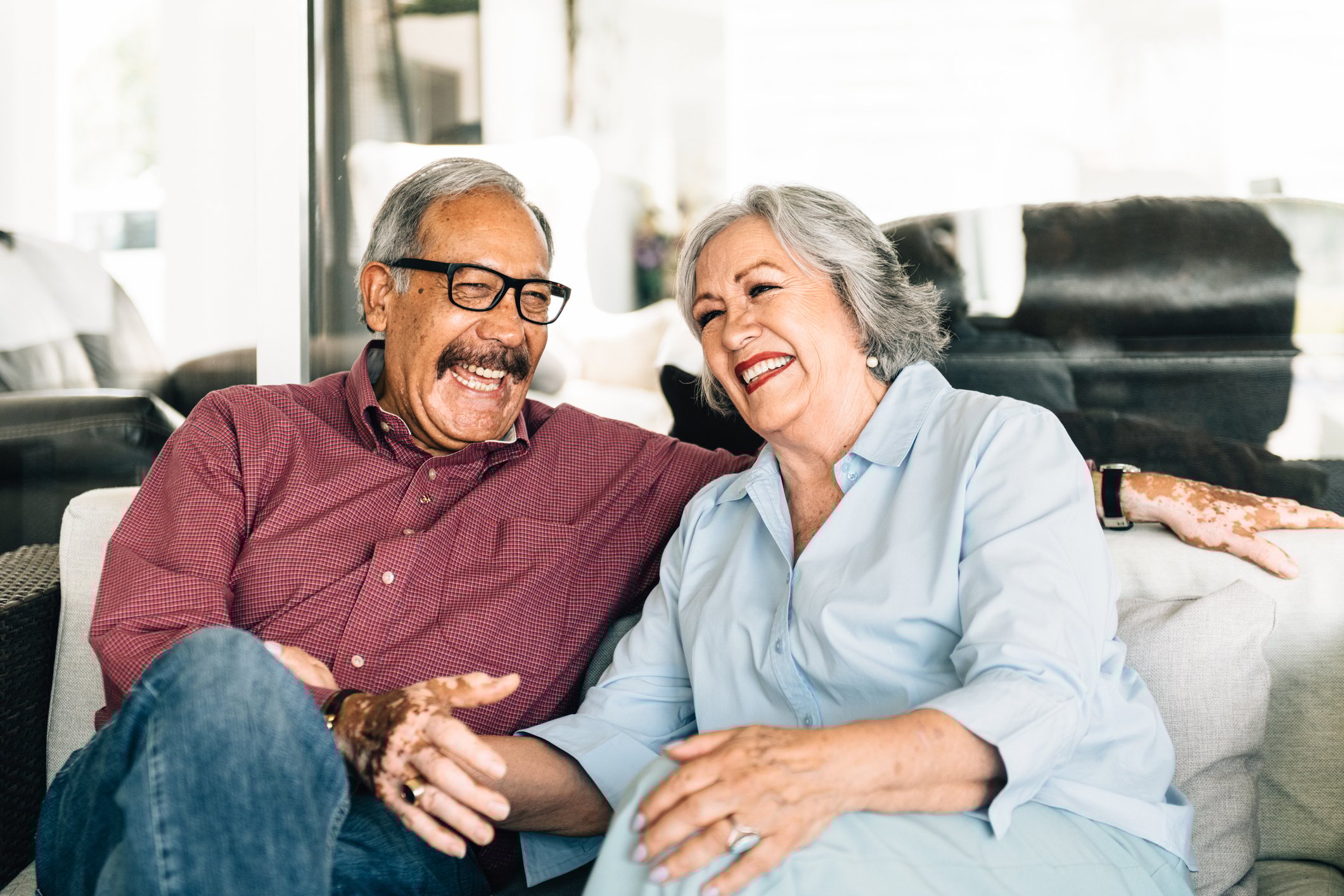 A silver-haired couple is on a couch, smiling broadly. 