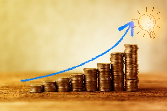 Stacks of coins moving higher like a rising stock chart with a light bulb at the end.