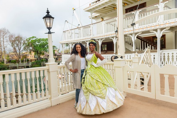 Tiana from "The Princess and the Frog" posing in front of Disney's signature riverboat with a park attendee.
