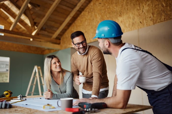 Three people stand around a table with a blueprint in a partially built building.