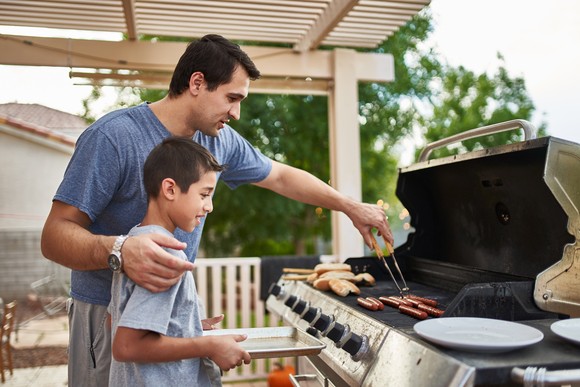 An adult and child smile while grilling hot dogs. 