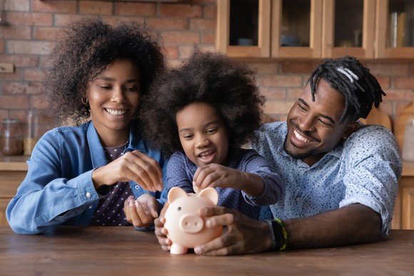 Two adults and a child sitting together at a table putting coins into a piggy bank.