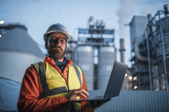 A worker wearing a safety helmet holds a laptop while standing in front of power plant towers