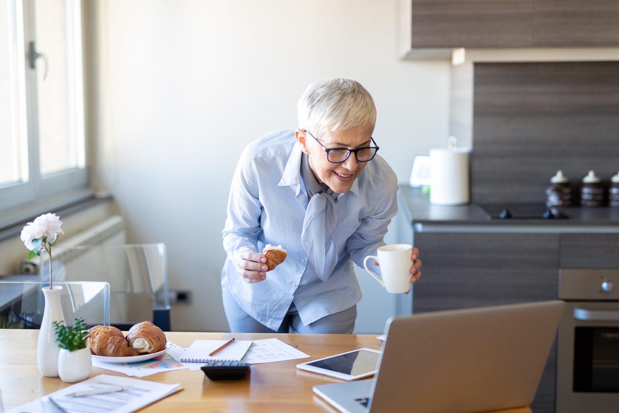 A woman stands in her kitchen holding a coffee cup while looking at her laptop sitting on a table with a smile on her face