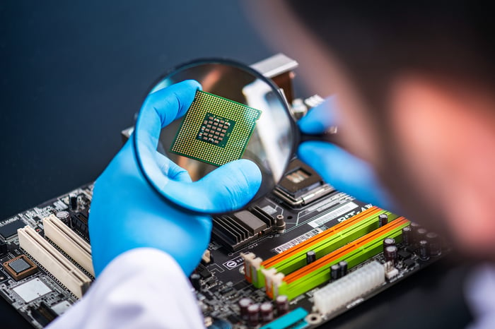 An engineer wearing rubber gloves looking at a microchip through a magnifying glass.