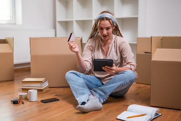A person surrounded by moving boxes in an empty home, using a credit card to buy something on a tablet computer.
