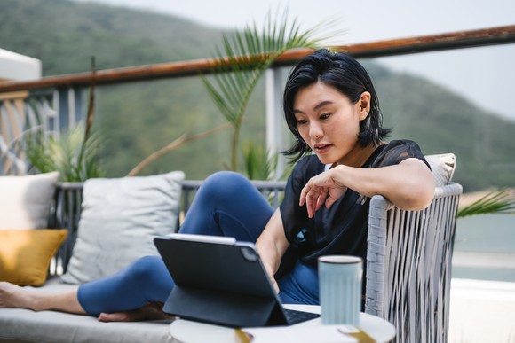 A person sitting on a couch on a balcony typing on a tablet. 