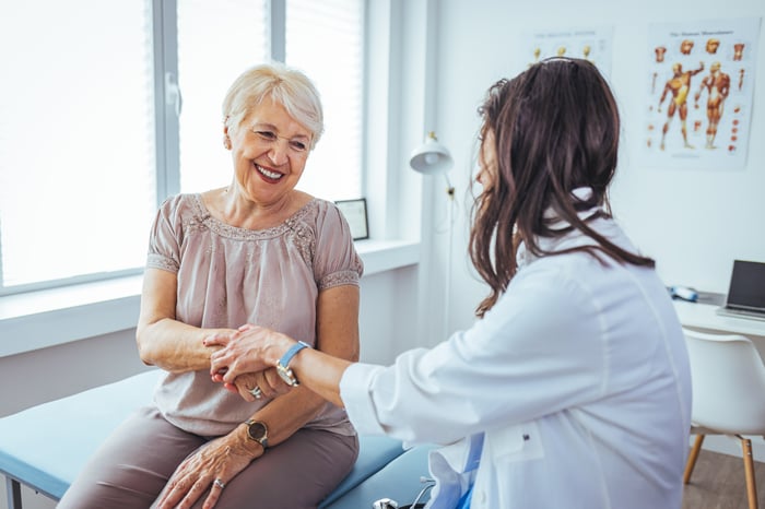 A person in a white coat holds the arm of a seated person.