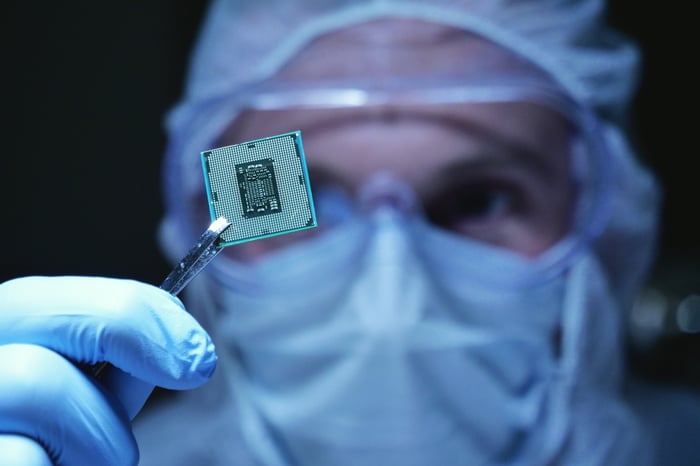 An engineer in a clear suit, mask, rubber gloves, and goggles holding up a microchip with tweezers. 