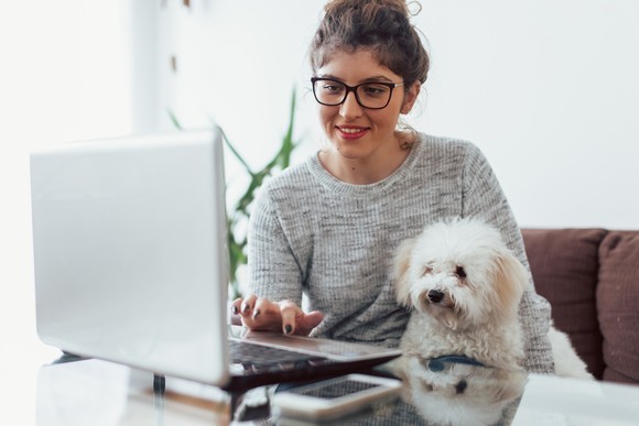 Smiling person typing on laptop, with dog in lap.