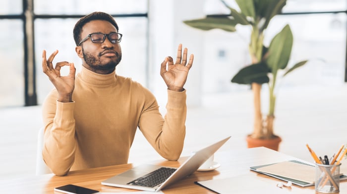 A man in front of his computer makes the ohm sign with his hands.